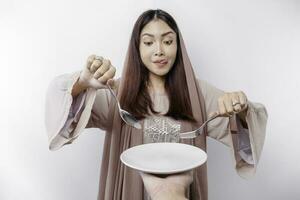 ein lächelnd asiatisch Muslim Frau ist Fasten und hungrig und halten und zeigen zu ein Teller foto