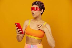 Frau Blogger Überraschung im ungewöhnlich tausendjährig Brille nehmen Selfies auf ihr Telefon Sport hell farbig Kleider gegen ein Orange Studio Hintergrund, kostenlos Raum foto