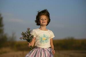 glücklich wenig Mädchen im das Wiese mit ein Strauß von Blumen. ein Kind auf ein schön Sommer- Feld mit Entwicklung Haar. foto