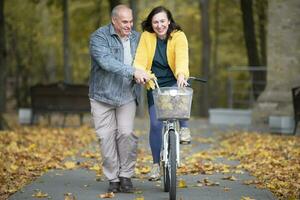 glücklich Mitte alt Paar mit Fahrrad im Herbst Park. Alten heiter Mann und Ehefrau auf ein gehen. foto