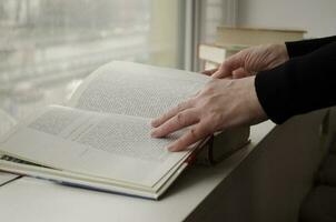 ein Person lesen Bücher in der Nähe von das Fenster. Hände wendet sich Über Buch Buchseite. foto