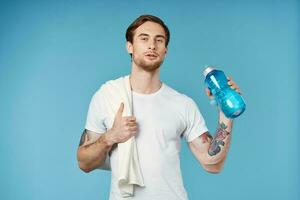 sportlich Mann zeigen Daumen oben Wasser Flasche Handtuch auf Schulter abgeschnitten Aussicht Blau Hintergrund foto