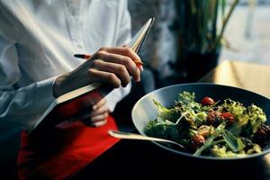 Frau mit Notizblock in der Nähe von das Fenster und Salat im ein Teller Tomaten frisch Gemüse foto