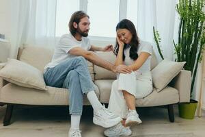 ein traurig asiatisch Frau Gespräche zu ein Mann im Tränen beim Zuhause Sitzung auf das Couch. jung Paar von anders Nationalitäten und Konflikt von Interesse im ein Paar foto