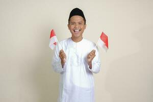 Porträt von attraktiv asiatisch Muslim Mann im Weiß Hemd mit Schädeldecke halten Indonesien Flagge während erziehen seine Faust, feiern Erfolg. isoliert Bild auf grau Hintergrund foto