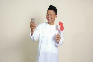 Porträt von attraktiv asiatisch Muslim Mann im Weiß Hemd mit Schädeldecke halten Indonesien Flagge während zeigen Handy, Mobiltelefon Telefon. isoliert Bild auf grau Hintergrund foto