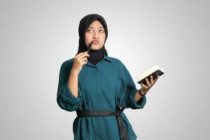 Porträt von aufgeregt asiatisch Muslim Frau mit Hijab Schreiben auf Hinweis Buch und Denken mit Stift auf kinn. Werbung Konzept. isoliert Bild auf Weiß Hintergrund foto