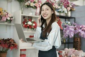 einer jung asiatisch weiblich Florist Eigentümer, Arbeiten mit Laptop, Verkauf Blumen- arrangieren, reden auf Handy, Mobiltelefon Telefon im bunt Blume Geschäft Geschäft mit ein schön Bündel von Blüten, und E-Commerce Geschäft. foto