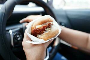 asiatisch Frau Treiber halt und Essen Hamburger im Auto, gefährlich und Risiko ein Unfall. foto