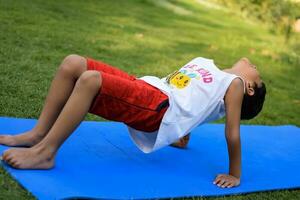 asiatisch Clever Kind tun Yoga Pose im das Gesellschaft Park draussen, Kinder- Yoga Pose. das wenig Junge tun Yoga und Meditation Übung. foto