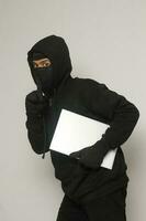 mysteriös Räuber Dieb Mann tragen schwarz Kapuzenpullover und Maske stehlen Laptop und schleichen aus. isoliert Bild auf grau Hintergrund foto
