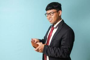 Porträt von jung asiatisch Geschäft Mann im beiläufig passen halten Leder Brieftasche und ziehen aus tausend Rupien während suchen beim Kamera lächelnd. isoliert Bild auf Blau Hintergrund foto