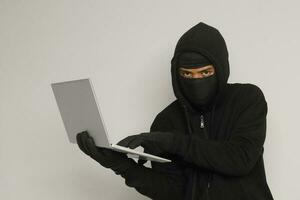 Porträt von mysteriös Mann tragen schwarz Kapuzenpullover und Maske tun hacken Aktivität auf Laptop, Hacker halten ein persönlich Computer. Cyber Sicherheit Konzept. isoliert Bild auf grau Hintergrund foto