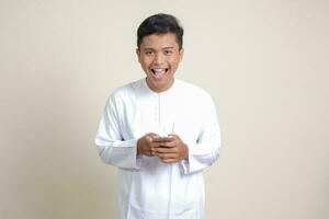 Porträt von attraktiv asiatisch Muslim Mann im Weiß Hemd halten Handy, Mobiltelefon Telefon mit lächelnd Ausdruck auf Gesicht. Werbung Konzept. isoliert Bild auf grau foto