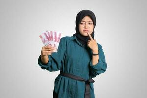 Porträt von verwirrt asiatisch Hijab Frau im beiläufig Outfit zeigen einer hundert tausend Rupiah während Denken mit Hand auf kinn. finanziell und Ersparnisse Konzept. isoliert Bild auf Weiß Hintergrund foto