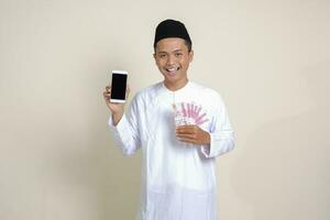 Porträt von attraktiv asiatisch Muslim Mann im Weiß Hemd mit Schädeldecke zeigen einer hundert tausend Rupiah während zeigen leer Bildschirm Handy, Mobiltelefon Telefon. finanziell und Einkaufen Konzept. isoliert Bild auf grau foto