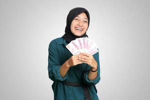 Porträt von aufgeregt asiatisch Hijab Frau im beiläufig Outfit zeigen einer hundert tausend Rupiah. finanziell und Ersparnisse Konzept. isoliert Bild auf Weiß Hintergrund foto