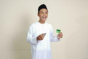 Porträt von attraktiv asiatisch Muslim Mann im Weiß Hemd mit Schädeldecke halten ein Handy, Mobiltelefon Telefon und präsentieren Anerkennung Karte. isoliert Bild auf grau Hintergrund foto