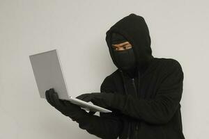 Porträt von mysteriös Mann tragen schwarz Kapuzenpullover und Maske tun hacken Aktivität auf Laptop, Hacker halten ein persönlich Computer. Cyber Sicherheit Konzept. isoliert Bild auf grau Hintergrund foto