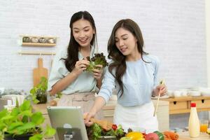 zwei jung asiatisch Frauen sind lesen Rezept von das Digital Tablette im das Küche. Frau haben Spaß mit gesund Essen Salat. foto