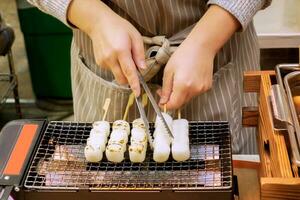 Hände von Koch mit ein Zangen Zange japanisch Snack auf das Grill zu bereiten zum Verkauf zu Kunden im das Markt. foto