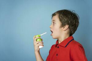 ein süß 5 Jahre alt kaukasisch Junge im ein rot T-Shirt Sprays ein Aerosol in seine Mund zu behandeln wund Kehle foto