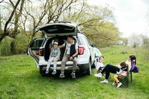 Familie mit drei Kinder gegen Auto öffnen Kofferraum auf Picknick. foto