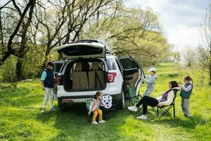 Mutter mit Kinder sitzen auf Stuhl gegen Auto öffnen Kofferraum auf Picknick. foto