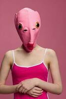 ein Frau tragen ein Rosa Fisch Kopf Maske auf Halloween steht und sieht aus beim das Kamera gegen ein Rosa Hintergrund. konzeptionelle Kunst Foto im ein verrückt Weg