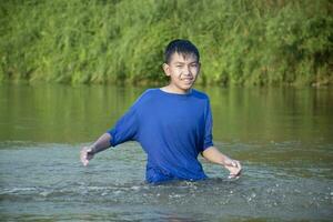 asiatisch Junge im Blau T-Shirt ist Ausgaben seine Freizeit durch Tauchen, Baden, werfen Felsen und fangen Fisch im das Fluss glücklich, Hobby und Glück von Kinder Konzept, im Bewegung. foto