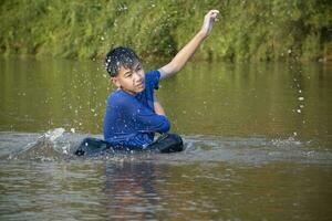 asiatisch Junge im Blau T-Shirt ist Ausgaben seine Freizeit durch Tauchen, Baden, werfen Felsen und fangen Fisch im das Fluss glücklich, Hobby und Glück von Kinder Konzept, im Bewegung. foto