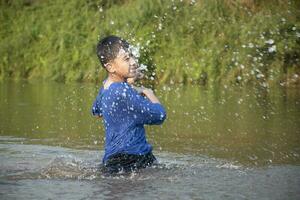 asiatisch Junge im Blau T-Shirt ist Ausgaben seine Freizeit durch Tauchen, Baden, werfen Felsen und fangen Fisch im das Fluss glücklich, Hobby und Glück von Kinder Konzept, im Bewegung. foto