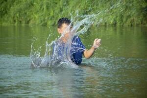 asiatisch Junge im Blau T-Shirt ist Ausgaben seine Freizeit durch Tauchen, Baden, werfen Felsen und fangen Fisch im das Fluss glücklich, Hobby und Glück von Kinder Konzept, im Bewegung. foto