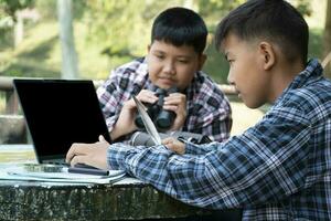 jung asiatisch Junge hält Vogel Feder und mit Laptop zu Geschäft und suchen Vögel Information mit seine Freund welche halten Fernglas hinter, selektiv Fokus auf Vorderseite Junge, Natur Studie Konzept. foto