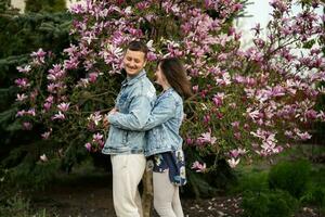 zärtlich Paar im Liebe tragen Jeans Jacke genießen nett Frühling Tag in der Nähe von Magnolie Blühen Baum. foto