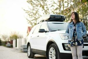 stilvoll Frau im Jeans Jacke Stehen in der Nähe von Weiß amerikanisch suv Auto mit Dach Gestell Kasten. foto