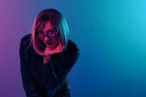 froh sexy schön blond Frau im Leder Jacke prickelnd Kleid modisch Sonnenbrille öffnen Mund berühren Kinn posieren isoliert im Farbe Licht Studio Hintergrund. Neon- Party Cyberpunk Konzept. Kopieren Raum foto