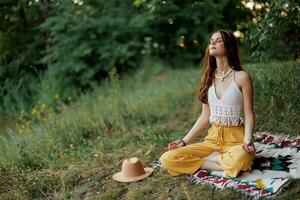 ein jung Hippie Frau meditiert im Natur im das Park, Sitzung im ein Lotus Position auf ihr bunt Plaid und genießen Harmonie mit das Welt im Öko-Kleidung foto