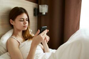 ziemlich Frau im das Morgen unter das Abdeckungen mit ein Handy, Mobiltelefon Telefon im ihr Hand foto