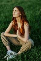 ein jung Frau im Sommer- Kleider Sitzung auf das Grün Gras tun Yoga und meditieren im Natur, ein Verbindung mit das Kosmos. das Konzept von Harmonie mit Körper und Natur, spirituell Wachstum foto