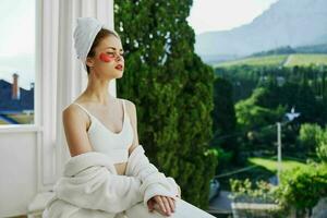 sinnlich und schön Frau mit ein Handtuch auf seine Kopf Patches auf seine Gesicht steht auf das Balkon perfekt sonnig Morgen foto