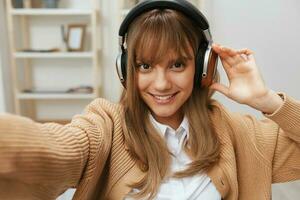 süß jung blond Schüler Dame Freiberufler im warm Sweatshirt im Kopfhörer Hör mal zu Favorit Lieder Sitzung im Sessel beim modern Zuhause Innere. Musik- Zeit entspannend cool Wiedergabeliste Konzept. breit Selfie Aussicht foto