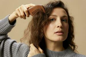 bezaubernd ziemlich lockig schön weiblich im grau beiläufig Sweatshirt mit Haarbürste Kamm gesund Locken posieren isoliert auf Über Beige Pastell- Hintergrund. problematisch widerspenstig beschädigt Haar Konzept. Kopieren Raum foto