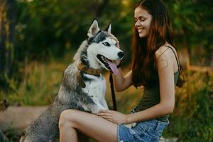 ein Frau mit ein heiser Rasse Hund lächelt und liebevoll Schlaganfälle ihr Geliebte Hund während Gehen im Natur im das Park im Herbst gegen das Hintergrund von Sonnenuntergang foto