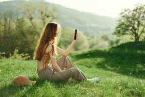 Frau Blogger sitzt auf das Grün Gras im ein Park und nimmt Bilder von Sie selber auf ihr Telefon gegen das Hintergrund von ein Sommer- Landschaft. jung Menschen Lebensstil und Besorgnis, Sorge zum das Umgebung foto