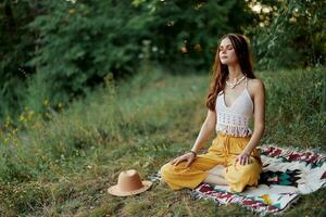 ein jung Hippie Frau meditiert im Natur im das Park, Sitzung im ein Lotus Position auf ihr bunt Plaid und genießen Harmonie mit das Welt im Öko-Kleidung foto