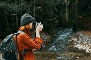 Frau Fotograf halten ein Kamera im Hand in der Nähe von das Fluss im das Berge und Wald im das Hintergrund foto