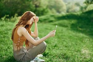 ein jung glücklich Frau Sitzung im das Park auf das Grün Gras im Natur mit ihr Telefon im ihr Hände im ihr beiläufig Kleider und chatten über Video Verknüpfung, das Konzept von gesund Erholung Meditation foto