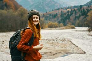 Frau im ein Sweatshirt Hut mit ein Rucksack ruhen in der Nähe von das Fluss im das Berge auf Natur Landschaft foto