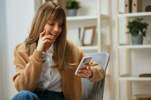 heiter jung blond Frau im warm Sweatshirt erhalten gut Nachrichten auf Studien durch Telefon berühren Wange Sitzung im Sessel beim heim. Pause von arbeiten, nehmen ein brechen, Sozial Medien im kostenlos Zeit Konzept. Kopieren Raum foto